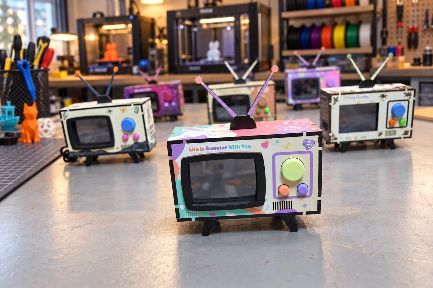 Several finished Mini TVs on a workbench with tools and 3D printers behind them